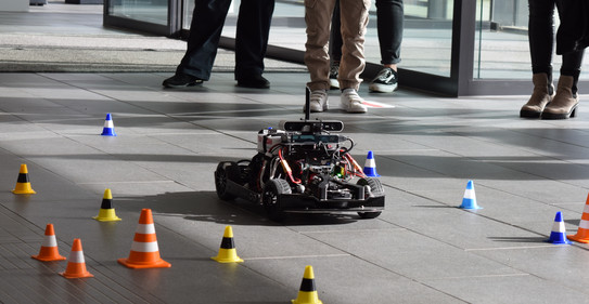 The AADC model car is driiving around a race track marked with traffic cones at the TU's 2023 Open House. Some people are observing.