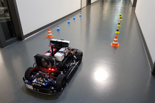 A model car driving down a hallway through a track marked by miniature traffic cones.