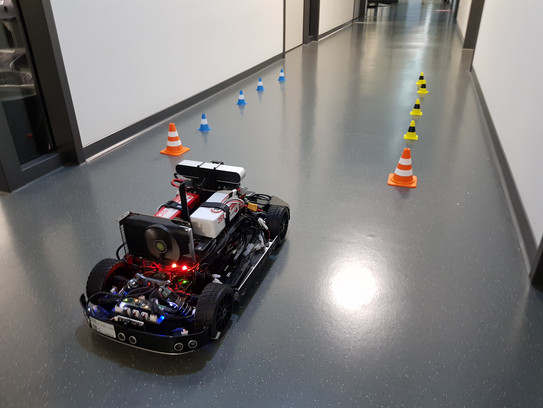 A model car driving down a hallway through a track marked by miniature traffic cones.